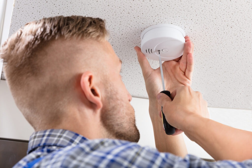 A person in a checkered shirt uses a screwdriver to install a smoke detector on a ceiling, highlighting home safety maintenance.